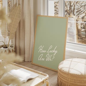 Pu&ograve; includere: Un poster verde salvia incorniciato con testo bianco che recita "How Lucky Are We?" Il poster &egrave; appoggiato su un pouf in vimini intrecciato in una stanza con una finestra e una pianta.