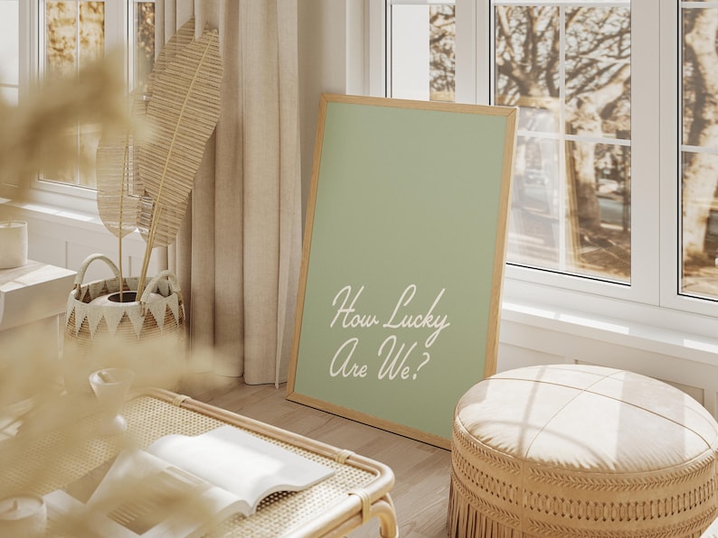 Pu&ograve; includere: Un poster verde salvia incorniciato con testo bianco che recita "How Lucky Are We?" Il poster &egrave; appoggiato su un pouf in vimini intrecciato in una stanza con una finestra e una pianta.