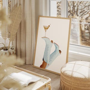 May include: A framed print of a pair of blue cowboy boots with a martini glass balanced on top. The background is a light beige color.