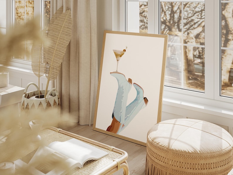 May include: A framed print of a pair of blue cowboy boots with a martini glass balanced on top. The background is a light beige color.