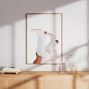 Op de afbeelding: Een ingelijste kunstdruk met een gestileerde illustratie van witte cowboylaarzen. De laarzen zijn afgebeeld tegen een witte achtergrond, met een houten frame. Het kunstwerk wordt getoond op een houten kast.