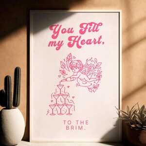 May include: Pink and white printable wall art with a graphic of a cupid pouring liquid into a stack of martini glasses. The text reads "You fill my heart, to the brim."