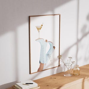 May include: A framed watercolor painting of a pair of blue cowboy boots with a martini glass on top. The boots are partially visible, with the martini glass resting on the top of the boots. The background is a light blue color.