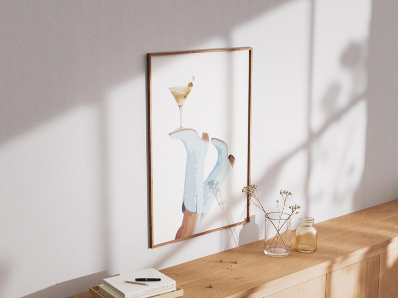 May include: A framed watercolor painting of a pair of blue cowboy boots with a martini glass on top. The boots are partially visible, with the martini glass resting on the top of the boots. The background is a light blue color.