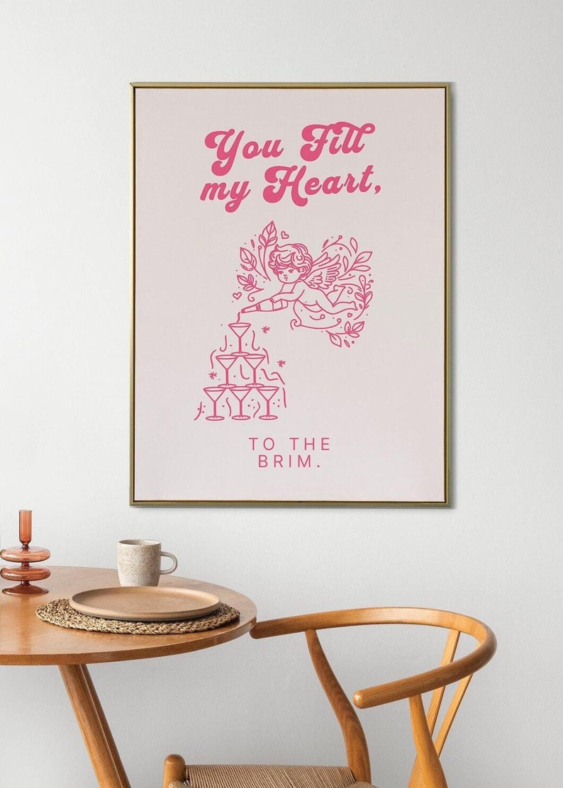 May include: Pink and white printable wall art with a gold frame. The art features a pink illustration of a cupid pouring champagne into a pyramid of glasses. The text reads "You fill my heart, to the brim."