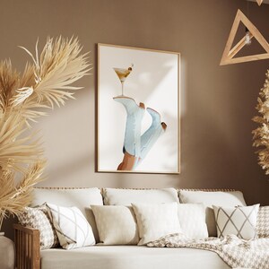 May include: A framed print of a pair of blue cowboy boots with a martini glass on top. The boots are on a white background. The print is hanging on a tan wall above a beige couch with pillows.