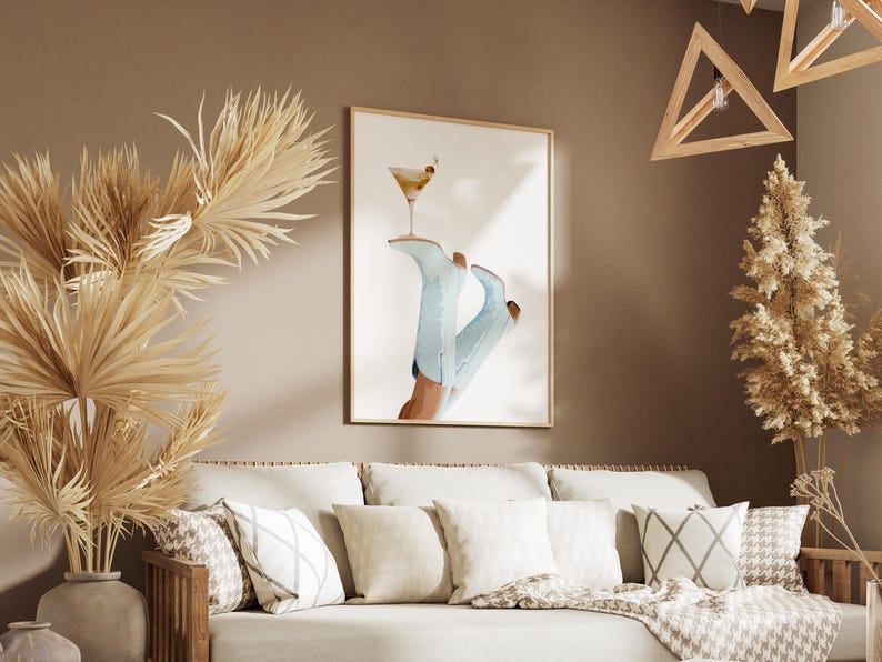 May include: A framed print of a pair of blue cowboy boots with a martini glass on top. The boots are on a white background. The print is hanging on a tan wall above a beige couch with pillows.