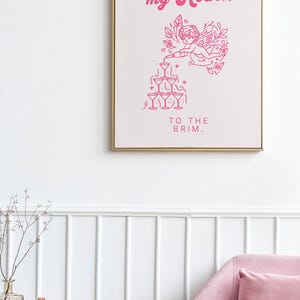May include: Pink and white framed print with a pink illustration of a cupid pouring champagne into a pyramid of glasses. The text reads "You fill my heart, to the brim."