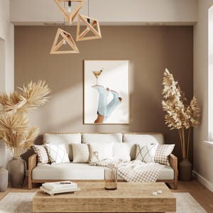 May include: A white couch with pillows and a throw blanket sits in a living room with a brown wall, a window, and a wooden coffee table. There is a dried palm plant in a brown pot and a dried grass plant in a brown pot. A framed print of a pair of blue boots with a cocktail glass hanging from the top of the boots is on the wall.