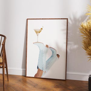 May include: A framed print of a pair of blue cowboy boots with a martini glass on top. The boots are tilted at an angle, and the martini glass is filled with a clear liquid and a green olive.