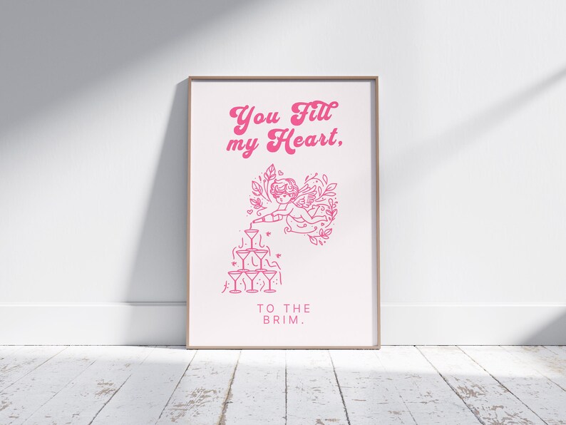 May include: Pink and white printable art print with a graphic of a cherub pouring champagne into a stack of glasses. The text reads "You fill my heart, to the brim."
