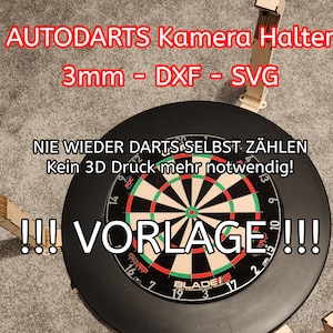 Może przedstawiać: Czarna tarcza do rzutków z czerwonym środkiem i białymi i zielonymi pierścieniami. Tarcza jest otoczona czterema drewnianymi uchwytami na kamerę, oznaczonymi jako "AUTODARTS Kamera Halter 3mm - DXF - SVG". Tekst "NIE WIEDER DARTS SELBST ZÄHLEN Kein 3D Druck mehr notwendig!" jest napisany nad tarczą. Tekst "!!! VORLAGE !!!" jest napisany pod tarczą.