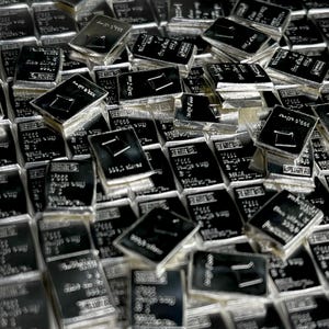 Peut inclure: Gros plan sur une pile de lingots d'argent. Chaque lingot est rectangulaire avec une surface sombre, possiblement noire, et présente du texte et un petit motif carré. Les lingots sont empilés, créant un motif dense et texturé.