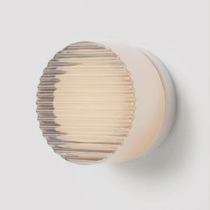 May include: A circular wall light in off-white with a ribbed, clear glass front. The light emits a warm glow. The design is modern and minimalist, suitable for various interior styles. The light is mounted on a white wall.