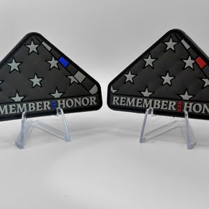May include: Two black, triangular patches resembling folded flags, each with white stars and the words "REMEMBER AND HONOR." One has a blue stripe, the other a red stripe. Both are displayed on clear stands.