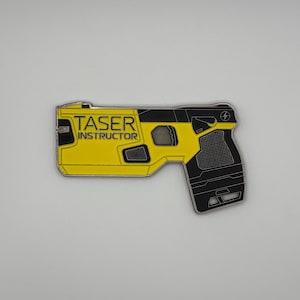 May include: A yellow and black Taser Instructor pin. The pin is shaped like a Taser gun with the words "TASER INSTRUCTOR" printed on the yellow portion. The pin has a black handle and a silver outline.