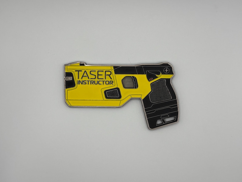 May include: A yellow and black Taser Instructor pin. The pin is shaped like a Taser gun with the words "TASER INSTRUCTOR" printed on the yellow portion. The pin has a black handle and a silver outline.