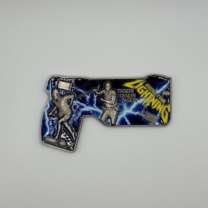 May include: A silver-colored metal coin in the shape of a gun, featuring two figures and lightning bolts against a blue background. The coin has the words "Ride the Lightning" and "Taser" on it.