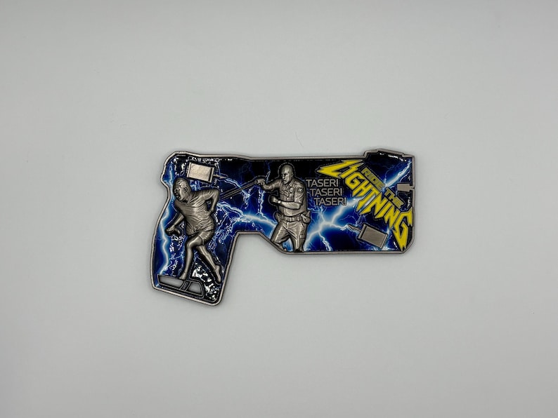 May include: A silver-colored metal coin in the shape of a gun, featuring two figures and lightning bolts against a blue background. The coin has the words "Ride the Lightning" and "Taser" on it.