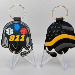 May include: Two keychains, each with a unique design. One features a black headset with emergency service symbols and the number "911" in yellow. The other displays a black and yellow American flag design. Both have a silver key ring.