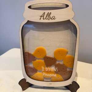 以下が含まれることがあります： 上部に「Alba」と刻まれた木製の瓶型貯金箱。瓶には黄色と茶色の形が入っています。「Je deviens propre」の文字が前面に見えます。銀行は木製のスタンドで支えられています。