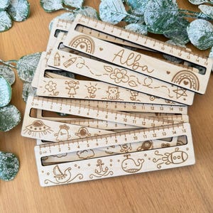 Puede incluir: Un conjunto de cinco reglas de madera con diferentes diseños y un nombre, "Alba", grabado en una de las reglas. Cada regla tiene un diseño diferente, incluyendo un arcoíris, un gato, una nave espacial y una ballena. Las reglas están hechas de madera marrón claro y tienen un fondo blanco.