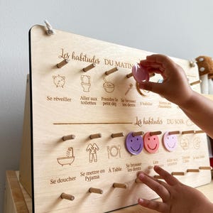 May include: Wooden morning and evening routine board with French text. The board features illustrations and pegs for tracking daily habits. A child's hand is placing a pink token on the board.