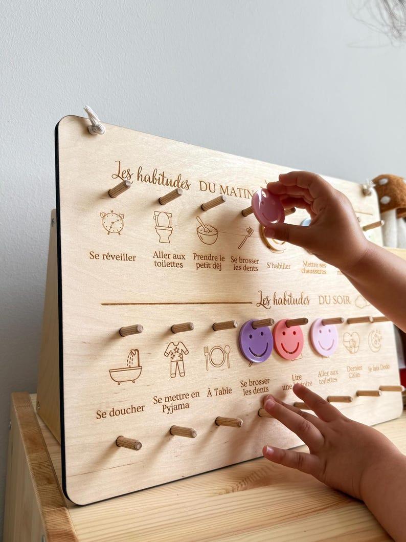 May include: Wooden morning and evening routine board with French text. The board features illustrations and pegs for tracking daily habits. A child's hand is placing a pink token on the board.