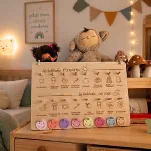 May include: A wooden morning and evening routine chart with colorful smiley face tokens. The chart has illustrations and French text. A plush monkey and other toys are on a wooden shelf. A framed picture with French text hangs on the wall.