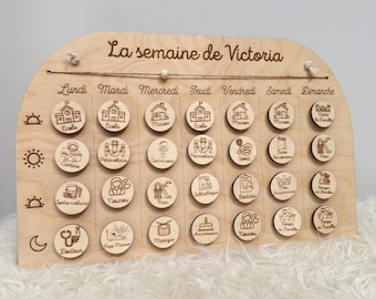 May include: Wooden weekly schedule board with engraved icons and text. The board is arched at the top and includes the text "La semaine de Victoria". Each day has circular wooden tokens with various activity icons.