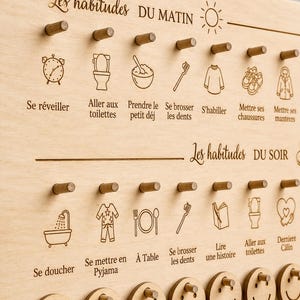 May include: Wooden morning and evening routine chart with engraved icons and text in French. Includes icons for waking up, brushing teeth, and getting dressed. Features smiley face tokens for completing tasks.