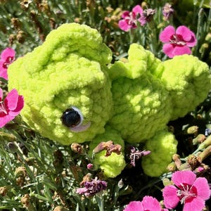 May include: A lime green crocheted plush toy shaped like a small creature with a textured, yarn-based surface. The figure features a single visible dark, glossy plastic eye with a white accent, rounded limbs, and a soft, rounded body structure.