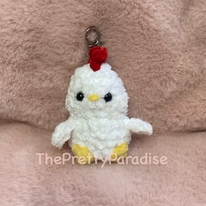 May include: A white, fluffy crochet chicken keychain with a red comb and yellow feet. The keychain has black eyes and a yellow beak. The text "ThePrettyParadise" is visible on the bottom of the image.