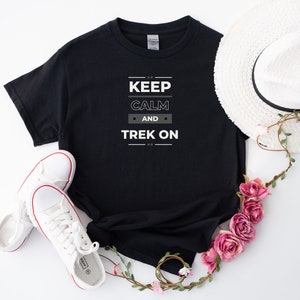 May include: Black t-shirt with white text that reads "Keep Calm and Trek On". The shirt is laid flat on a white surface with white trainers and a white sun hat with pink flowers.