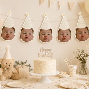 May include: A birthday party setting with a cake, balloons, and decorations. The cake has a "Happy Birthday" cake topper. The wall is decorated with baby faces wearing party hats. A teddy bear and gift box are also present.