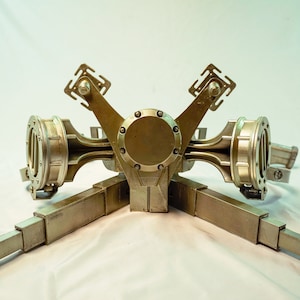 May include: A gold-colored metal object with four arms extending out from a central hub. Each arm has a square-shaped piece at the end. The object is on a white background.