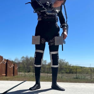May include: A black bodysuit worn with a metallic silver exoskeleton harness featuring chest plating, shoulder attachments, and thigh-mounted rectangular panels. The outfit includes silver knee guards and a handheld silver prop pistol accessory.