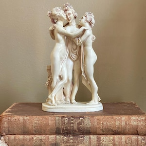 May include: A cream-colored sculpture depicting three figures embracing. The classical-style statue is set atop a base and is placed on top of two antique books with gold lettering. The figures are nude, with detailed facial features and flowing hair.
