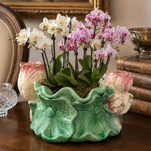 May include: A ceramic planter shaped like green lily pads with pink lotus flower accents, containing multiple stems of white and light purple orchids. The orchids are supported by thin black stakes and are planted in a bed of green moss.