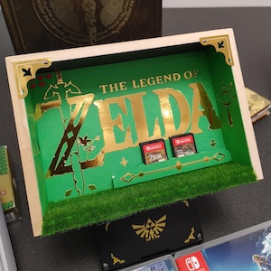 Può includere: Una scatola ombra in legno con "The Legend of Zelda" in lettere dorate su sfondo verde. Include due cartucce di gioco e dettagli decorativi dorati. La base ha una consistenza erbosa.