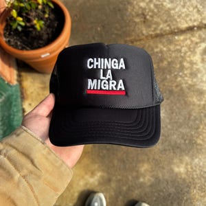 May include: Black trucker hat with the text "CHINGA LA MIGRA" in white, with a red stripe. The hat has a mesh back and a curved brim. The hat is being held up by a person.