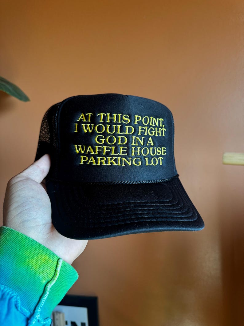 May include: Black trucker hat with the embroidered text "AT THIS POINT, I WOULD FIGHT GOD IN A WAFFLE HOUSE PARKING LOT" in yellow. The hat has a mesh back and a curved brim.
