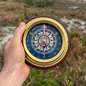 May include: A round compass with a dark blue face, gold-colored trim, and a brown wooden frame. The compass needle points north. The face has gold-colored markings and the words "TRUEST NORTH".