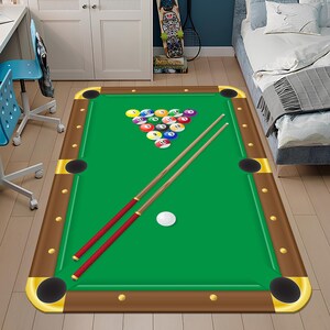 May include: A rectangular rug designed to look like a pool table. It has a green playing surface, brown borders, and corner pockets. A rack of numbered billiard balls and two pool cues are present.