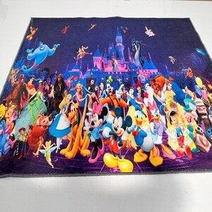 May include: A vibrant blanket displaying a collage of Disney characters in front of a castle. The blanket features characters from various Disney films, including Mickey Mouse, Donald Duck, and others, set against a dark purple backdrop.