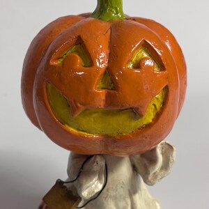 May include: A ceramic Halloween figurine of a ghost with a jack-o'-lantern head. The ghost is wearing a white sheet and holding a small pumpkin in its hand.