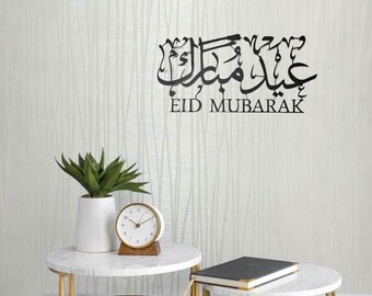 Könnte beinhalten: Schwarze Wanddekoration aus Metall mit arabischer Kalligraphie, die "Eid Mubarak" lautet. Die Dekoration ist an einer weißen Wand mit einem weißen Marmortisch und einer Uhr angebracht.