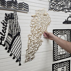 K&ouml;nnte beinhalten: Eine Wanddekoration aus schwarzen und goldenen Papierschnitten. Die Ausschnitte haben die Form eines Keffiyehs, einer Moschee und arabischer Kalligraphie. Die Kalligraphie hat die Form des Landes Libanon.