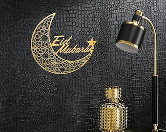 Könnte beinhalten: Goldener Halbmond aus Metall mit einem filigranen Design und den Worten "Eid Mubarak" darauf. Der Mond hängt an einer schwarzen, strukturierten Wand mit einer goldenen Lampe und einer goldenen Flasche auf einem Tisch darunter.