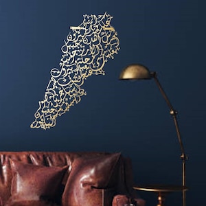 K&ouml;nnte beinhalten: Ein goldfarbener Wandaufkleber in Form des Libanon, bestehend aus arabischer Kalligraphie. Der Aufkleber befindet sich an einer dunkelblauen Wand, mit einem braunen Ledersofa und einer goldenen Lampe im Hintergrund.